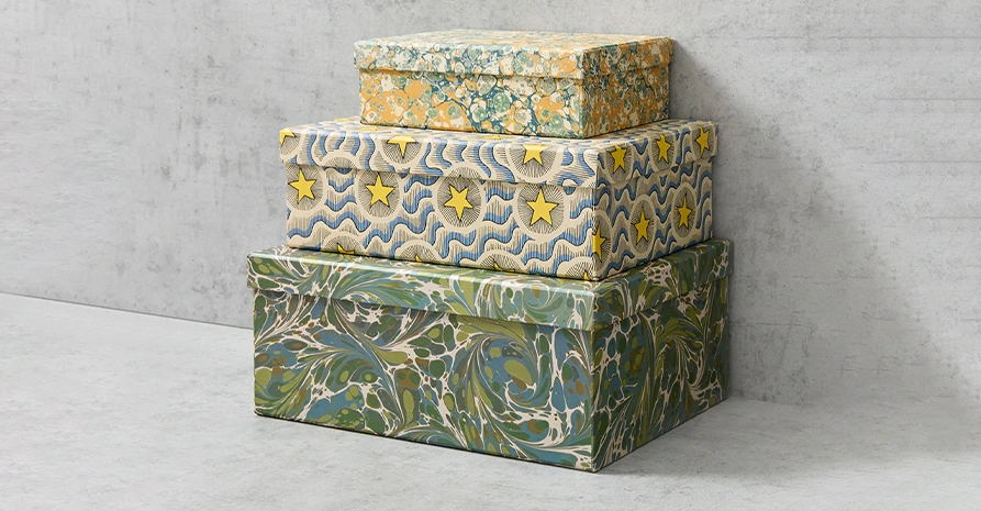 Three stacked decorative boxes with intricate patterns: the top box features a floral design, the middle has wavy lines and yellow stars, and the bottom showcases a green and blue marbled pattern. They are against a gray backdrop.